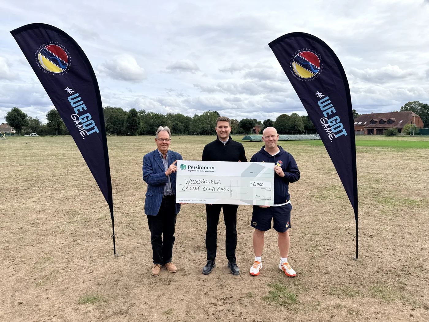 Councillor George Cowcher, Spencer Walters of Persimmon Homes South Midlands and Ross Owen of Wellesbourne Cricket Club with the £6,000 cheque, between two Wellesbourne Cricket Club Girls banners